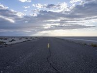 the empty road stretches out into the distance towards the sunset over water, sand, and cloudy skies