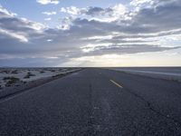 the empty road stretches out into the distance towards the sunset over water, sand, and cloudy skies