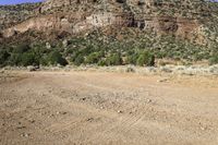 a dirt field with mountains and trees in the background in a desert area with some rocks and grass in the foreground