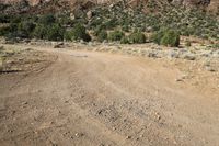 a dirt field with mountains and trees in the background in a desert area with some rocks and grass in the foreground