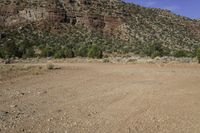 a dirt field with mountains and trees in the background in a desert area with some rocks and grass in the foreground