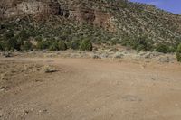 a dirt field with mountains and trees in the background in a desert area with some rocks and grass in the foreground