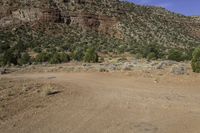 a dirt field with mountains and trees in the background in a desert area with some rocks and grass in the foreground