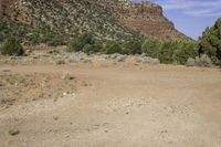 a dirt field with mountains and trees in the background in a desert area with some rocks and grass in the foreground