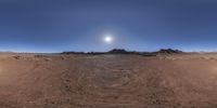 a wide panoramic photograph of a desert landscape with sun behind it and mountain peaks in the distance