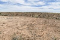 a very large empty dry field by a blue sky and clouds above it and some vegetation