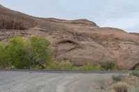 a motorcycle is traveling down a desert road near rocky hills, trees and bushes on the roadside