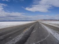 an empty asphalt road leading towards the desert mountains and blue skies in the distance, snow on it