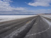 an empty asphalt road leading towards the desert mountains and blue skies in the distance, snow on it