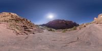 a panoramic view of a narrow river in the desert with red rocks, shrubs and a blue sky