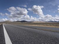 an empty open road runs through the desert under a sky filled with clouds, and hills in the background