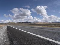 an empty open road runs through the desert under a sky filled with clouds, and hills in the background