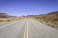 the road in desert is wide and empty with sparse brush, mountains and sand on either side