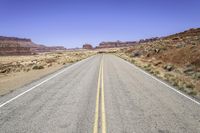 the road in desert is wide and empty with sparse brush, mountains and sand on either side