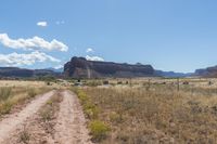 a dirt road next to mountains in the distance with sky and clouds above it on a sunny day