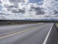 an empty highway surrounded by grass on a cloudy day with some clouds above it and power lines and mountains in the distance