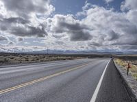 an empty highway surrounded by grass on a cloudy day with some clouds above it and power lines and mountains in the distance