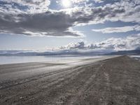 a gravel road stretching into the distance with water, mountains and clouds in the background