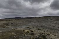 a dirt road and dirt hills during a cloudy day with dark clouds above them, in a remote area