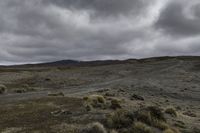 a dirt road and dirt hills during a cloudy day with dark clouds above them, in a remote area