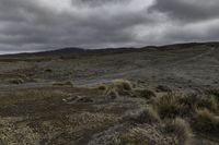 a dirt road and dirt hills during a cloudy day with dark clouds above them, in a remote area