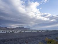 an empty street with rocks and bushes around it and mountains on the other side with water under a cloudy sky