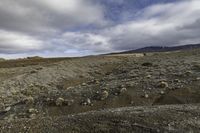 a lone person walking across a dirt field near mountains under a cloudy sky and mountains in the distance