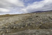 a lone person walking across a dirt field near mountains under a cloudy sky and mountains in the distance