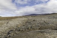 a lone person walking across a dirt field near mountains under a cloudy sky and mountains in the distance