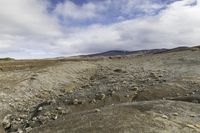 a lone person walking across a dirt field near mountains under a cloudy sky and mountains in the distance