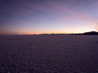 an empty snow field with the sun setting in the distance and mountains in the distance