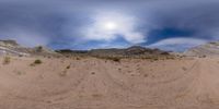 a panoramic view of mountains, dirt and grass against a cloudy blue sky