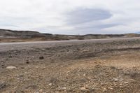 an empty desert road in the middle of nowhere land with grass, gravel, rocks and trees in the distance