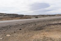 an empty desert road in the middle of nowhere land with grass, gravel, rocks and trees in the distance