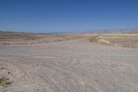 the dirt road winds through a dry desert plain against a blue sky and mountain range in the distance