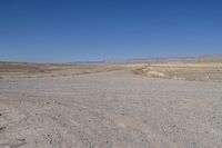 the dirt road winds through a dry desert plain against a blue sky and mountain range in the distance