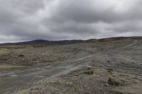 the dirt road leads to a cloudy sky above mountains and rocks and dirt, there is a lone motorcycle parked on a flat field