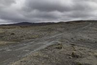 the dirt road leads to a cloudy sky above mountains and rocks and dirt, there is a lone motorcycle parked on a flat field