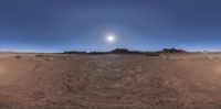 a wide panoramic photograph of a desert landscape with sun behind it and mountain peaks in the distance