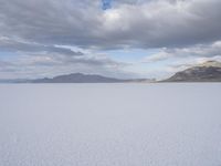 an open plain covered in white snow under a cloudy sky and mountain range behind the horizon