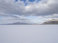 an open plain covered in white snow under a cloudy sky and mountain range behind the horizon