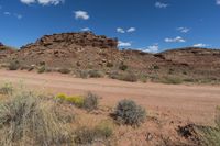 a dirt road near a field and hills in the desert with fluffy white clouds in the sky