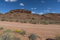 a dirt road near a field and hills in the desert with fluffy white clouds in the sky