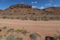 a dirt road near a field and hills in the desert with fluffy white clouds in the sky