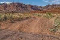 the dirt road goes through an arid, rocky terrain area with tall, flat red mountains in the distance