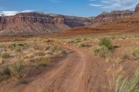 the dirt road goes through an arid, rocky terrain area with tall, flat red mountains in the distance