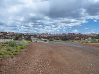 a dirt road in a barren landscape under some clouds, near some vegetation, and a road in the distance