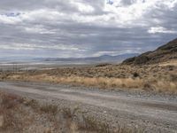 a dirt road through the desert on a cloudy day with mountains in the distance near a hill