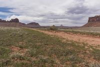 a dirt road with bushes on the side and tall rock formations in the background under a cloudy blue sky