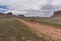 a dirt road with bushes on the side and tall rock formations in the background under a cloudy blue sky